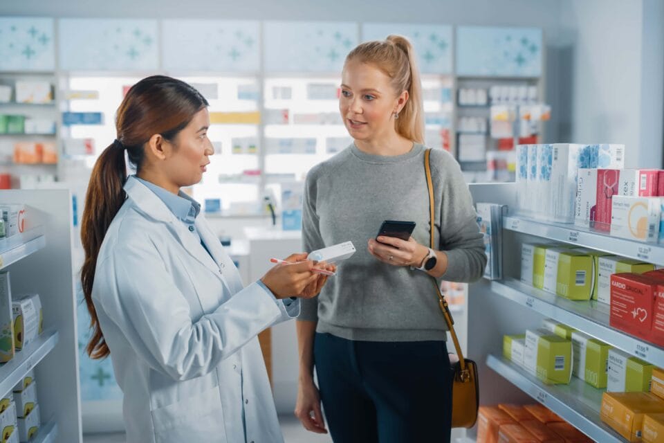 Pharmacy Drugstore: Caucasian Woman Chooses to Buy Medicine Professional Asian Pharmacist Advising, Consulting, Recommending Customer the Best Option. Modern Pharma Store Health Care Products